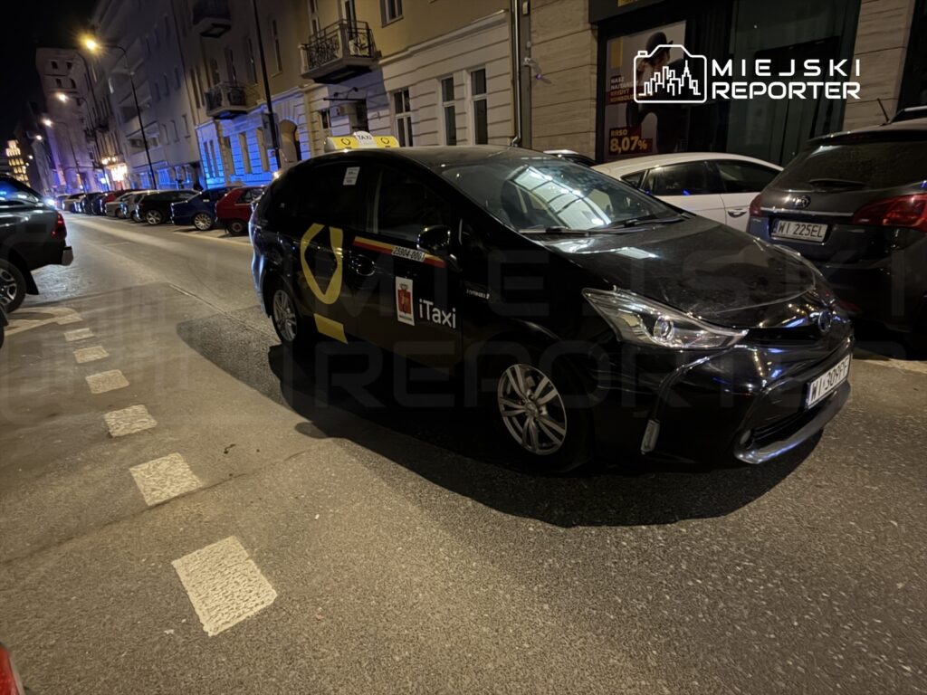 Czarny samochód taxi zaparkowany na ulicy nocą, obok innych pojazdów, w oświetlonym miejskim otoczeniu.