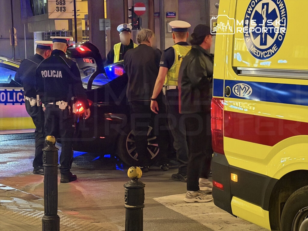 Grupa policjantów sprawdzająca wnętrze samochodu w pobliżu karetki pogotowia na ulicy.