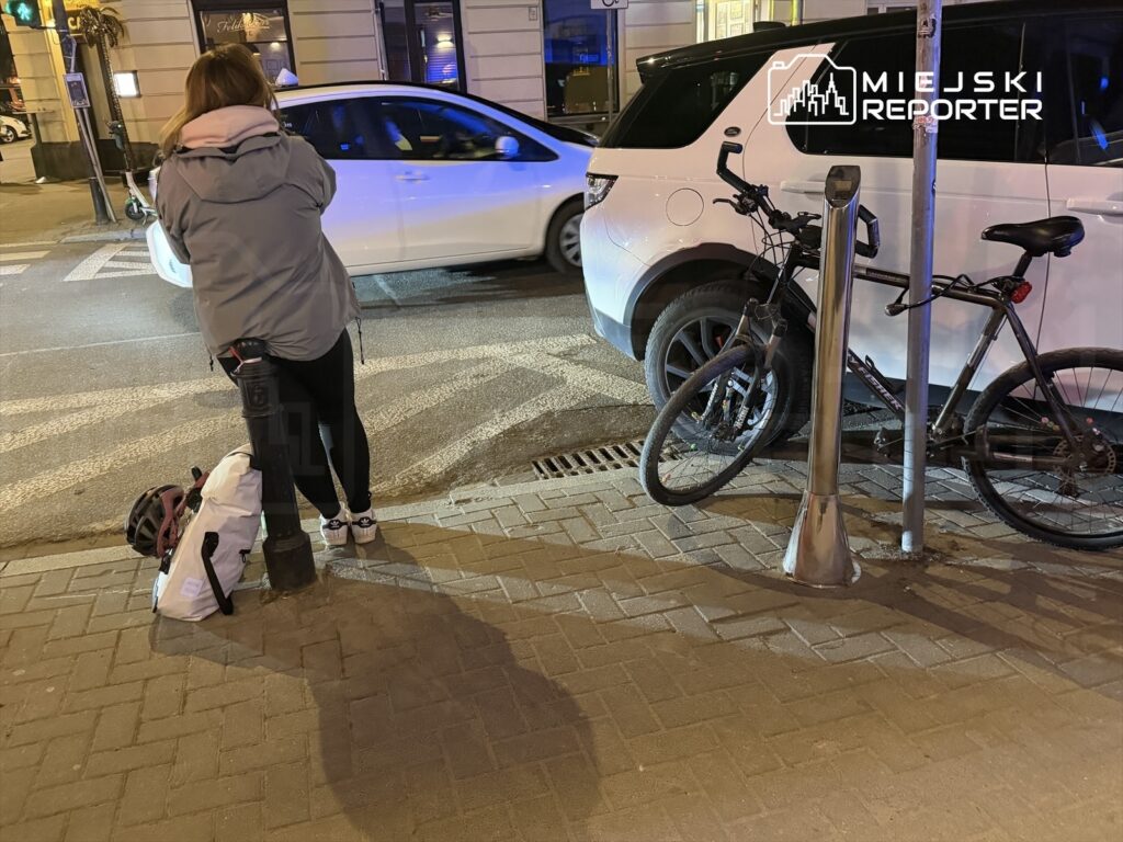 Kobieta w szarej kurtce stoi na chodniku obok zaparkowanego białego SUV-a, trzymając torbę i kask.