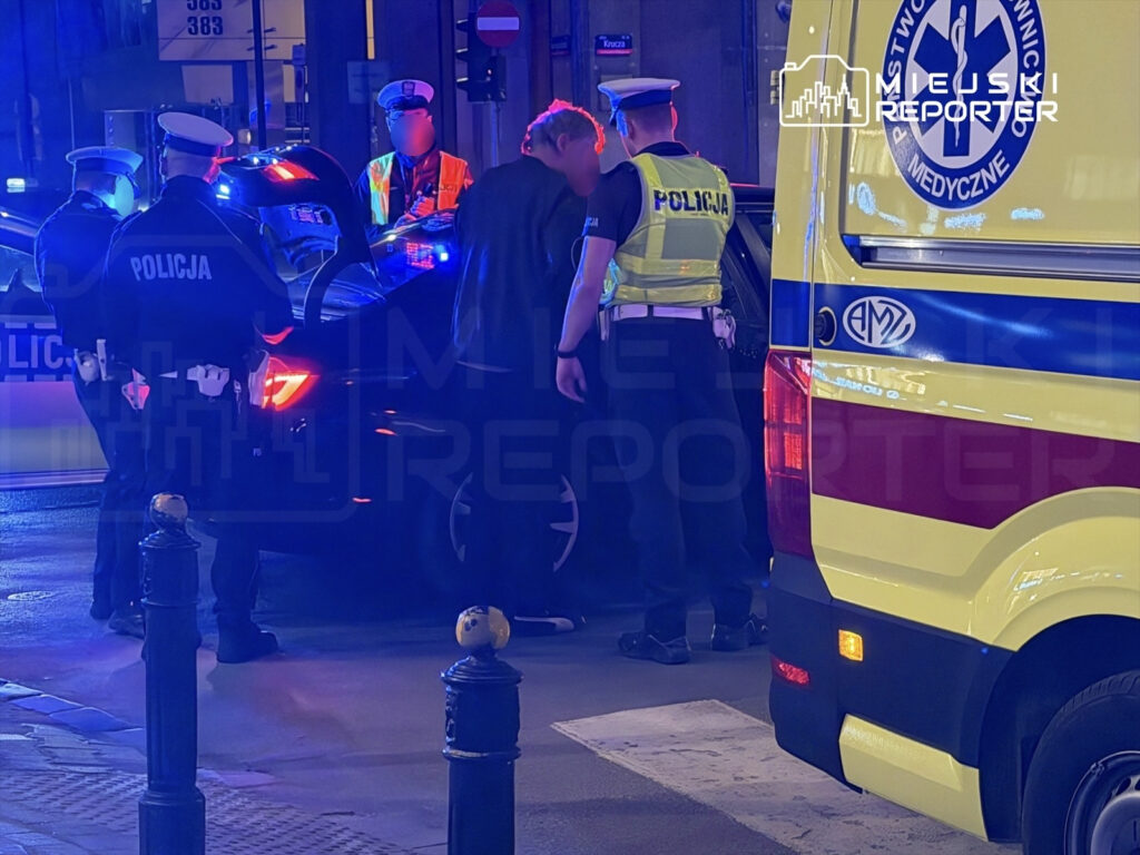 Grupa policjantów w mundurach interweniuje przy samochodzie, obok stoi ambulans z włączonymi sygnałami świetlnymi.
