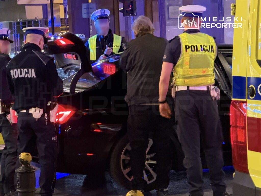 Policjanci sprawdzają zawartość bagażnika czarnego samochodu na ulicy, w tle widać radiowóz z włączonymi sygnałami świetlnymi.