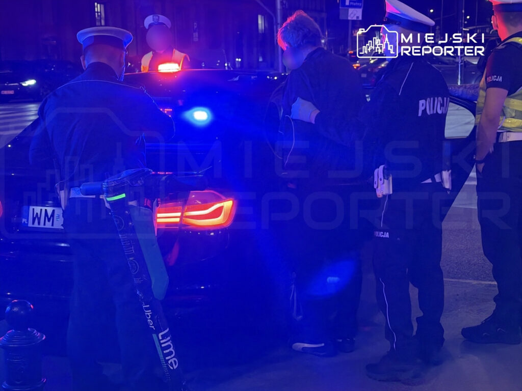 Policjanci w mundurach kontrolują osobę przy zaparkowanym samochodzie, otoczeni niebieskimi światłami policyjnymi.
