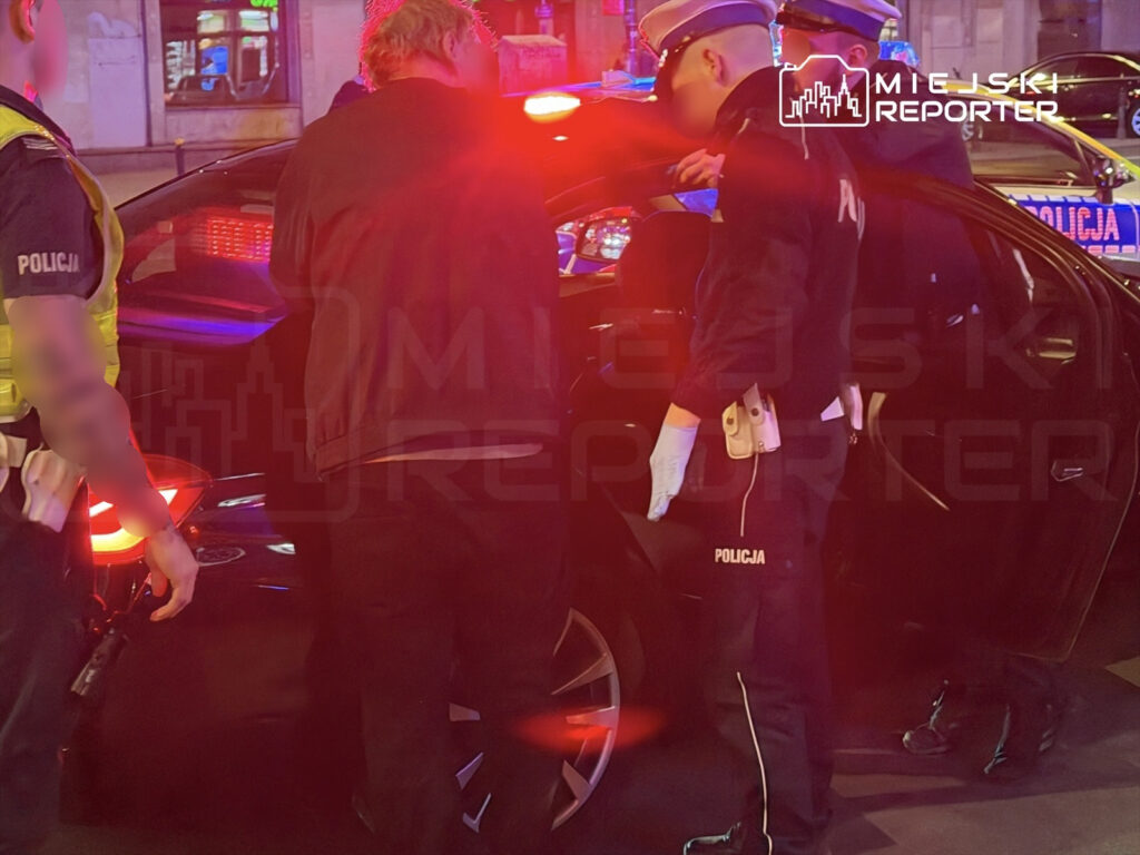 Policjanci kontrolują mężczyznę przy czarnym samochodzie w nocy, w tle migające światła radiowozów.