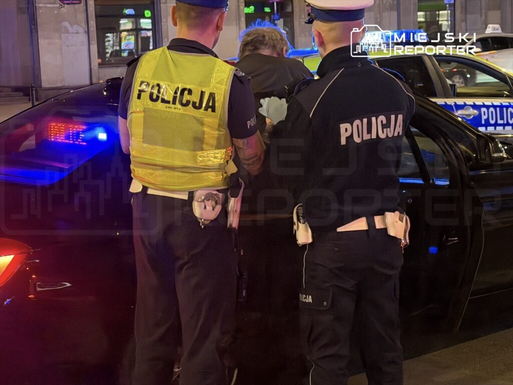 Dwóch policjantów w kamizelkach odblaskowych zatrzymuje mężczyznę w nocy przy radiowozie, w tle migają niebieskie światła.