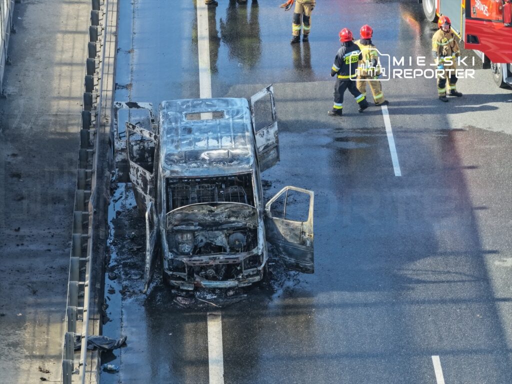 Spalony pojazd osobowy na pasie ruchu, obok strażacy prowadzący działania ratunkowe.