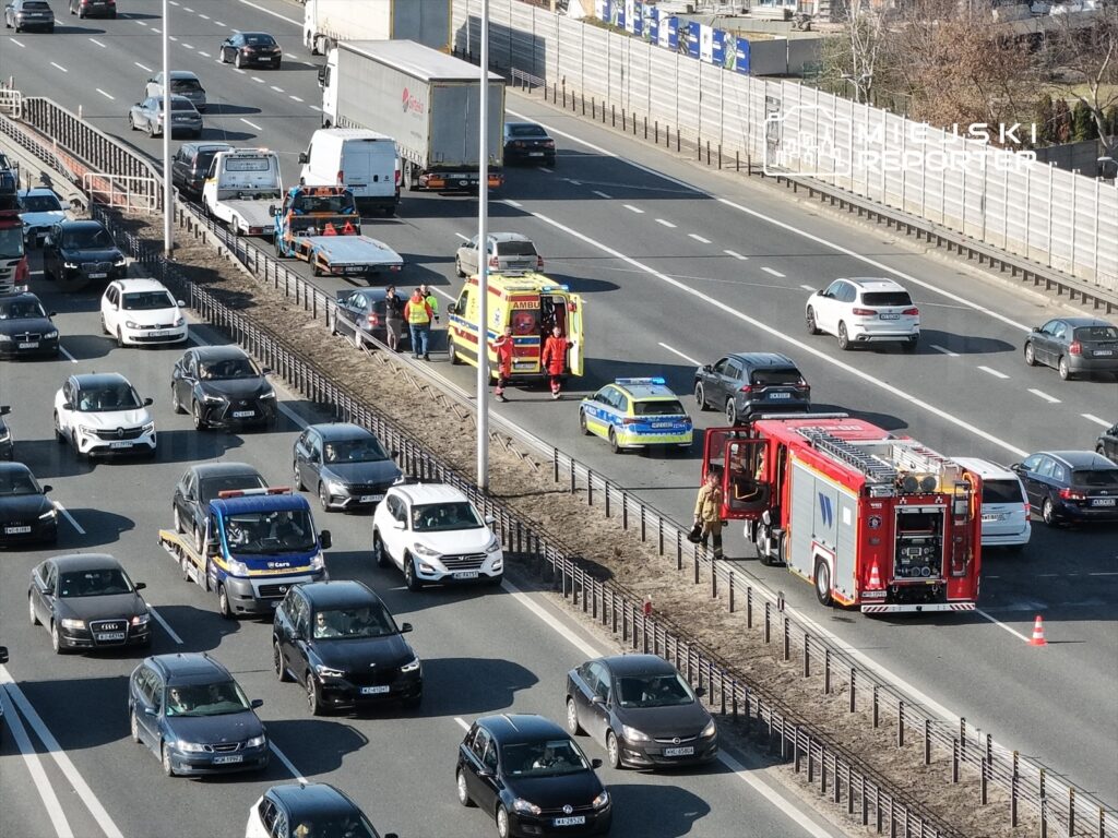Na zatłoczonej drodze ratownicy oraz strażacy interweniują przy wypadku drogowym. W tle widać pojazdy w ruchu.