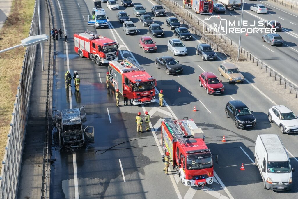 Na autostradzie, strażacy gaszą pożar spalonego samochodu, obok stoją dwa wozów strażackich, a w tle ruchliwy ruch drogowy.