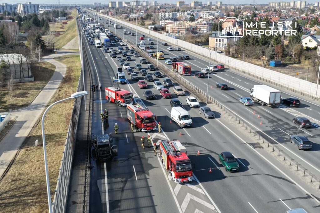 Na autostradzie zatrzymane pojazdy, w tym dwa wozy strażackie, a obok nich strażacy zabezpieczają miejsce po pożarze samochodu.