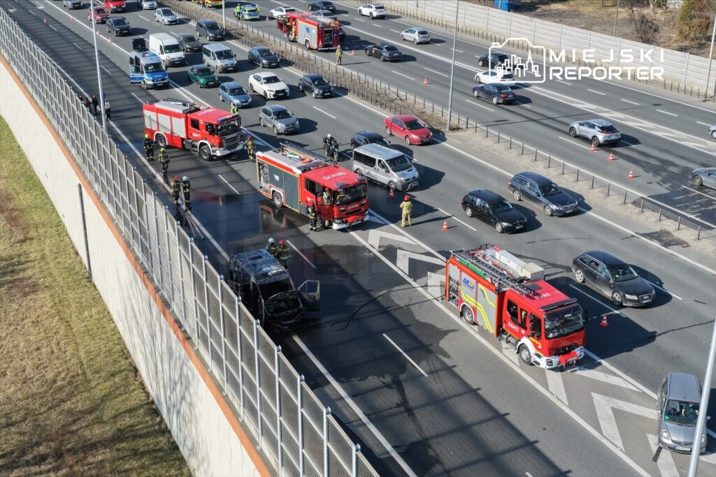 Wielu strażaków pracuje przy gaszeniu pożaru uszkodzonego pojazdu na drodze szybkiego ruchu.