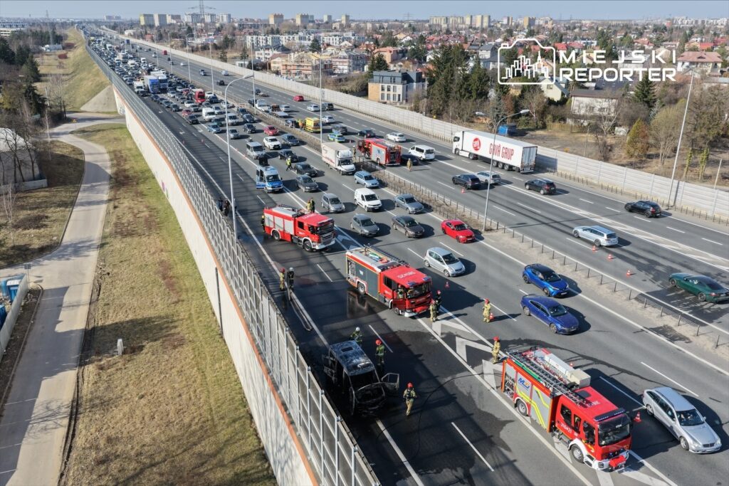 Na jezdni stoi samochód pożarowy, a strażacy gaszą płonący pojazd, obok zator drogowy.