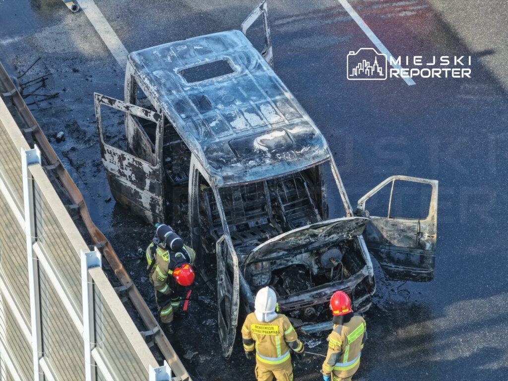 Strażacy w specjalistycznym sprzęcie badają spalone auto na drodze, obok widoczne ślady pożaru.