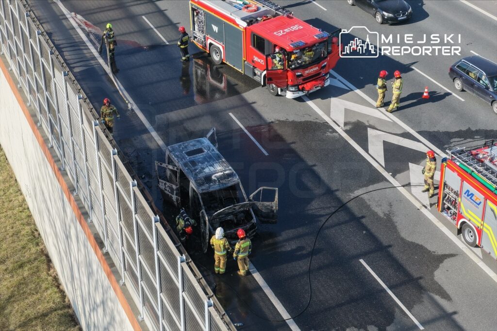 Strażacy prowadzą działania ratunkowe przy palącym się pojeździe na autostradzie, otoczonym przez pojazdy służb.