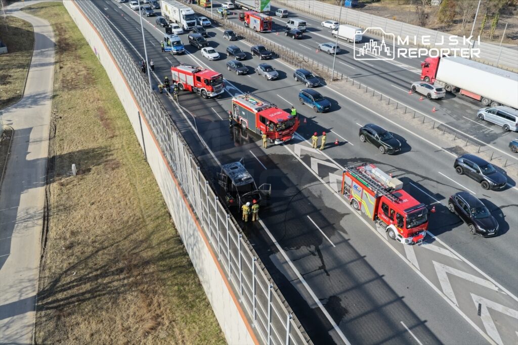 Na jezdni zatrzymane są pojazdy, a strażacy gaszą ogień w uszkodzonym samochodzie. W tle widać ruch uliczny.