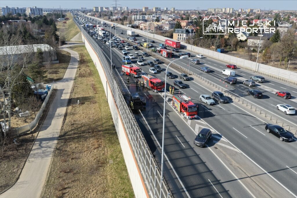 Na autostradzie zatrzymane pojazdy, obok strażacy gasić pożar samochodu, w tle widoczne budynki mieszkalne.
