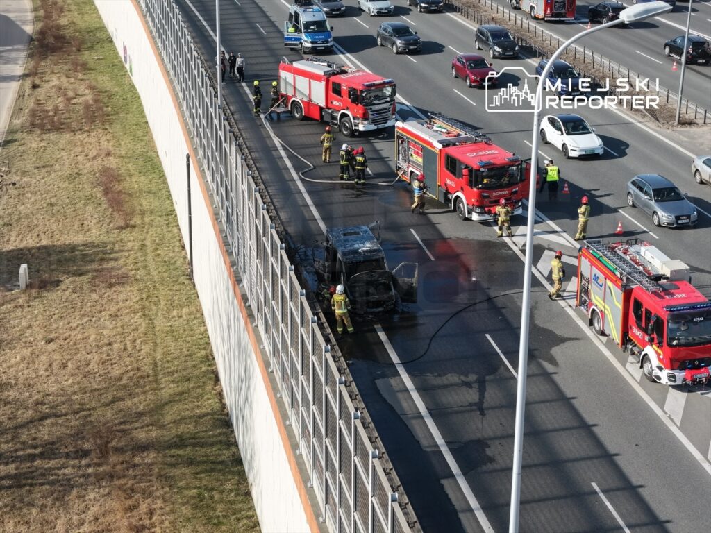 Strażacy gaszą płonący samochód na autostradzie, obok stoją pojazdy ratunkowe i zablokowany ruch.