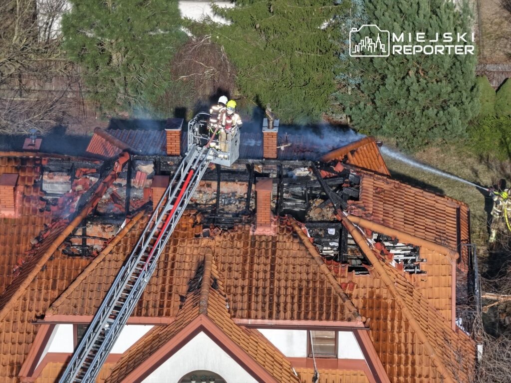 Strażacy gaszą pożar na dachu budynku, używając podnośnika, otoczeni przez zniszczoną konstrukcję.