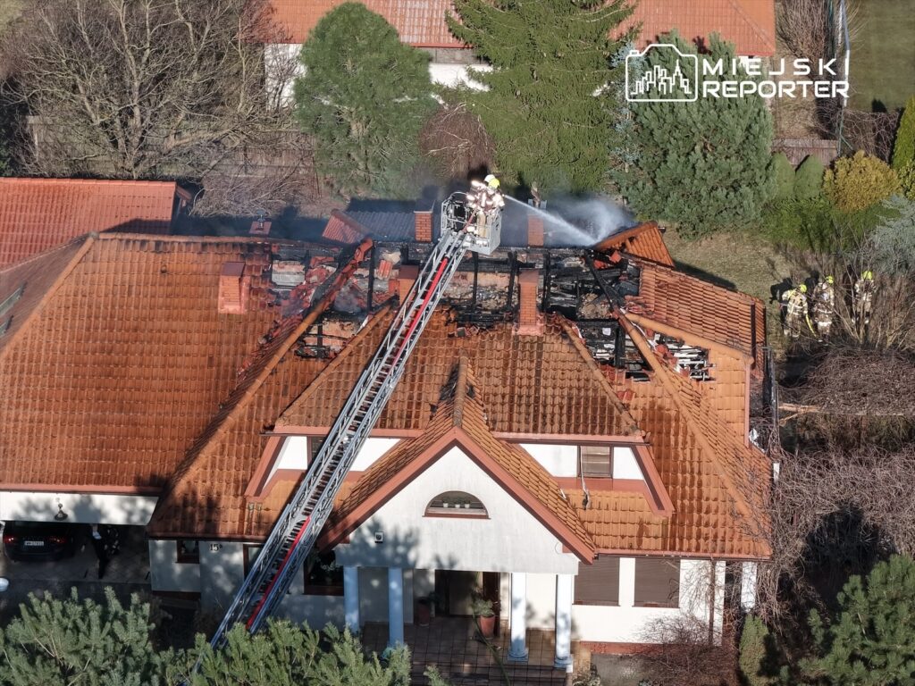 Strażacy gaszą pożar dachu domu jednorodzinnego, zniszczonego przez ogień, w otoczeniu zieleni.