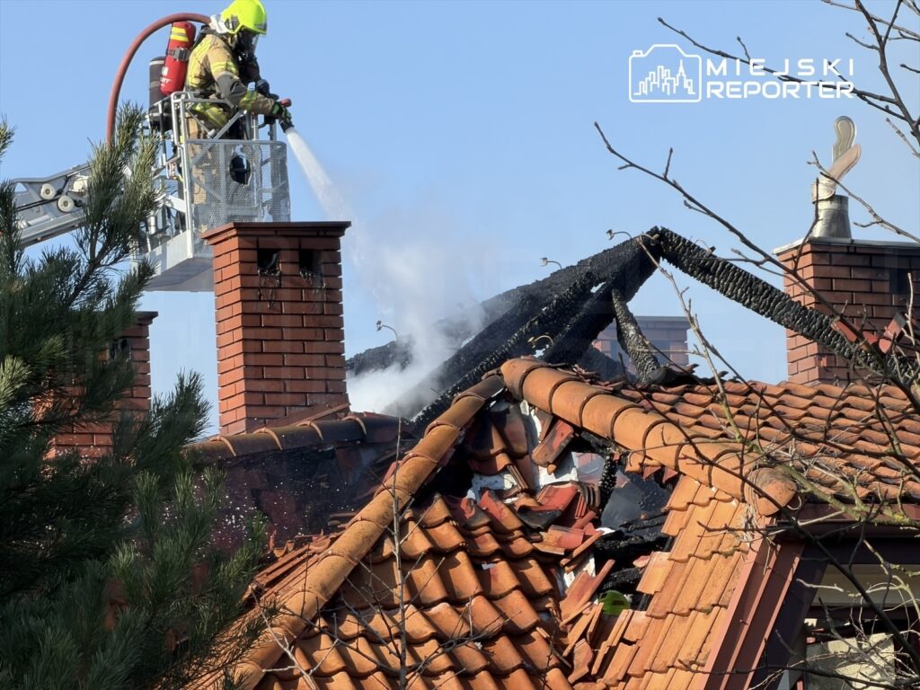 Strażak w kasku i mundurze, używający węża strażackiego z podnośnika, gasi pożar na dachu budynku mieszkalnego.