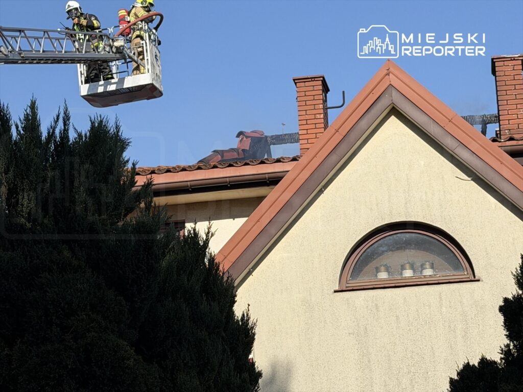 Strażacy w kaskach pracują na podnośniku, naprawiając dach budynku mieszkalnego w słoneczny dzień.