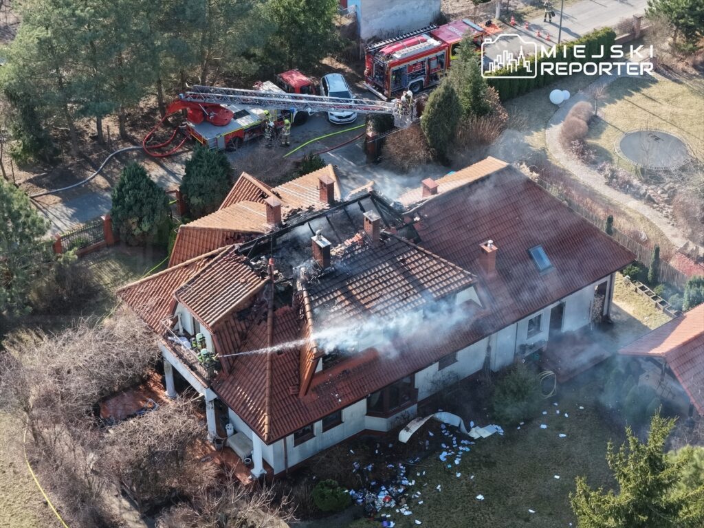 Strażacy gaszą pożar dachu domu jednorodzinnego, z którego wydobywa się dym, w otoczeniu zieleni.