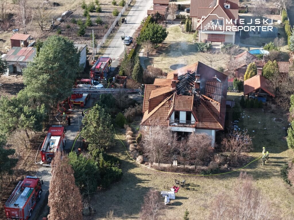 Strażacy pracują nad ugaszeniem pożaru w domu jednorodzinnym z uszkodzonym dachem, otoczeni przez pojazdy strażackie.