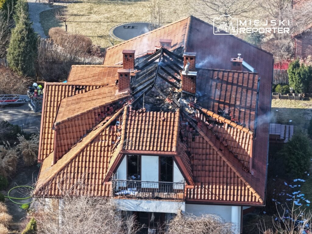 Dach domu jednorodzinnego zniszczony przez pożar, z widocznymi płomieniami i dymem, obok strażacy na drabinie.