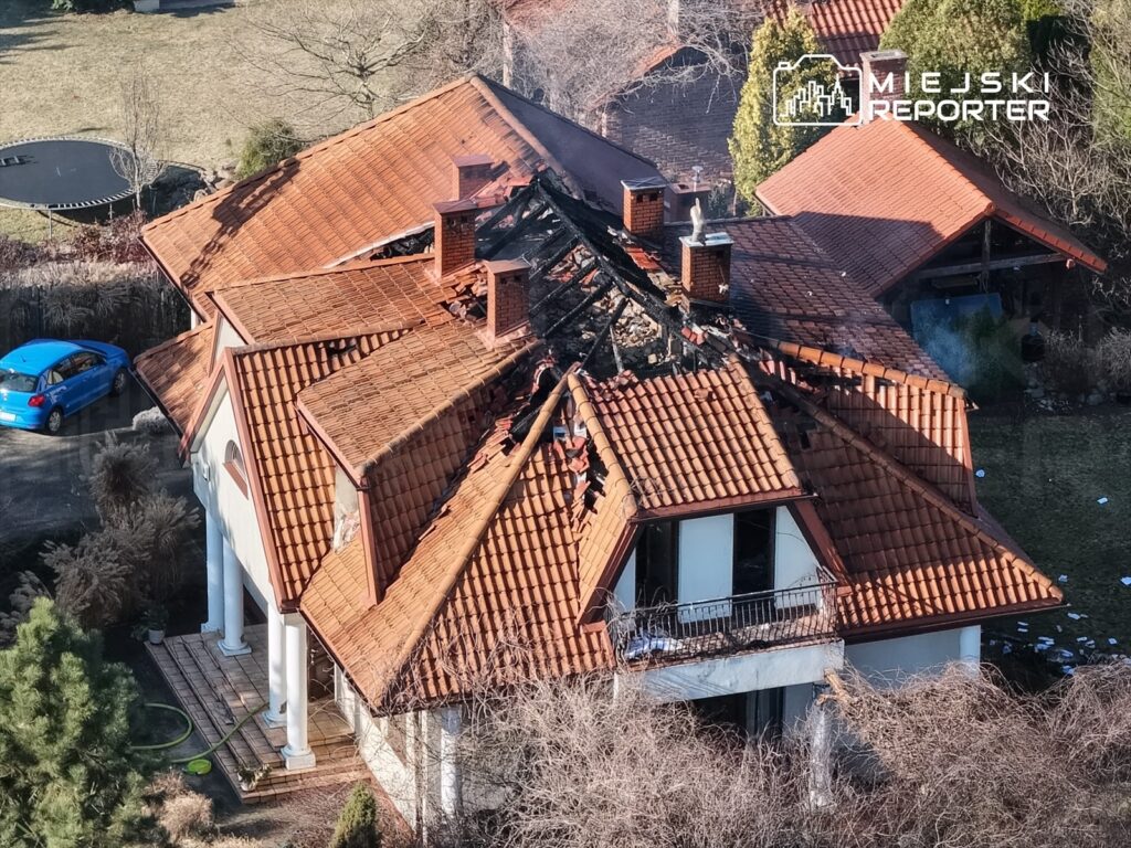 Dom z uszkodzonym dachem, z którego uwalnia się dym, w otoczeniu ogrodu i samochodu na podjeździe.