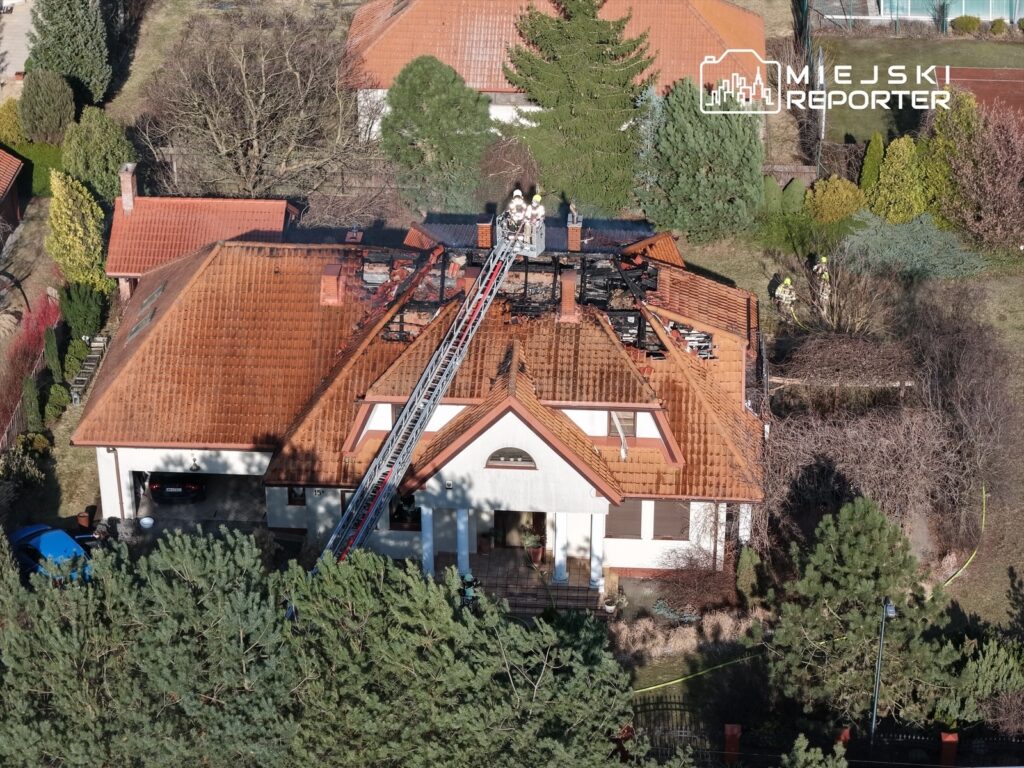 Strażacy gaszą pożar na dachu budynku jednorodzinnego, otoczonego zielenią, z widoczną zniszczeniem w górnej części.