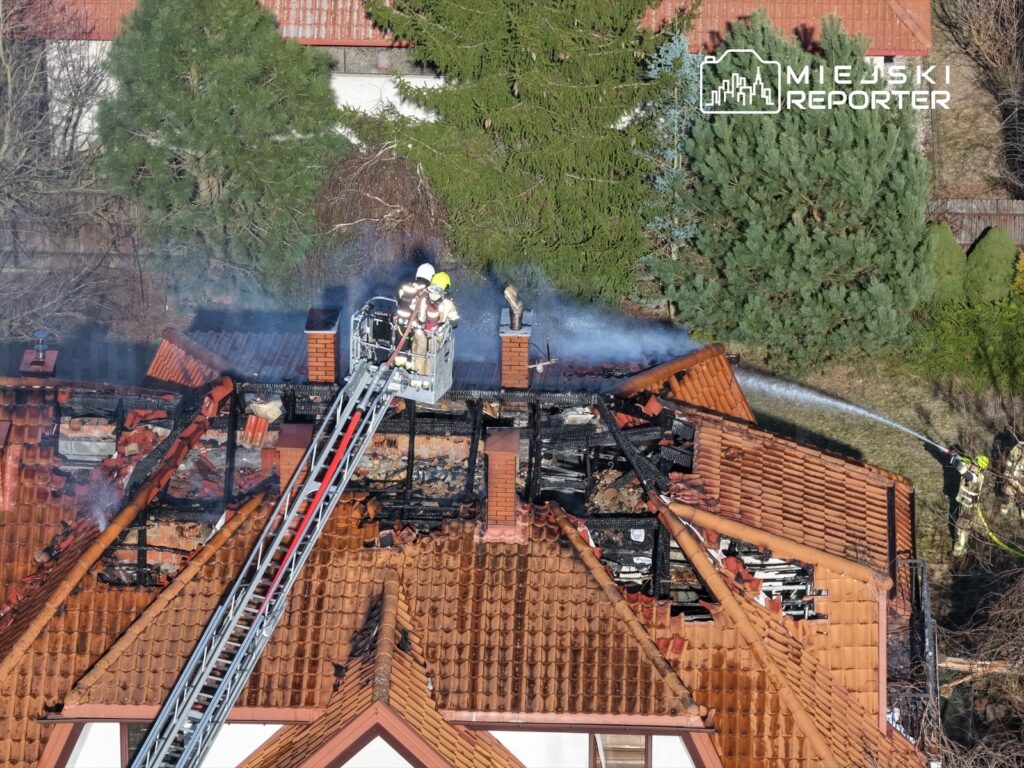 Strażacy gaszą pożar na dachu budynku, używając węża strażackiego z drabiny. W tle widoczne zniszczenia.