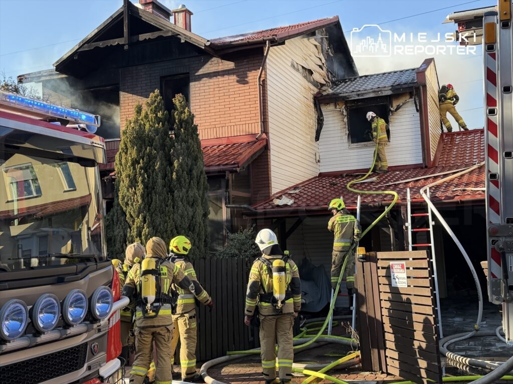 Strażacy w specjalistycznych strojach gaszą pożar na dachu budynku jednorodzinnego, otoczeni sprzętem gaśniczym.