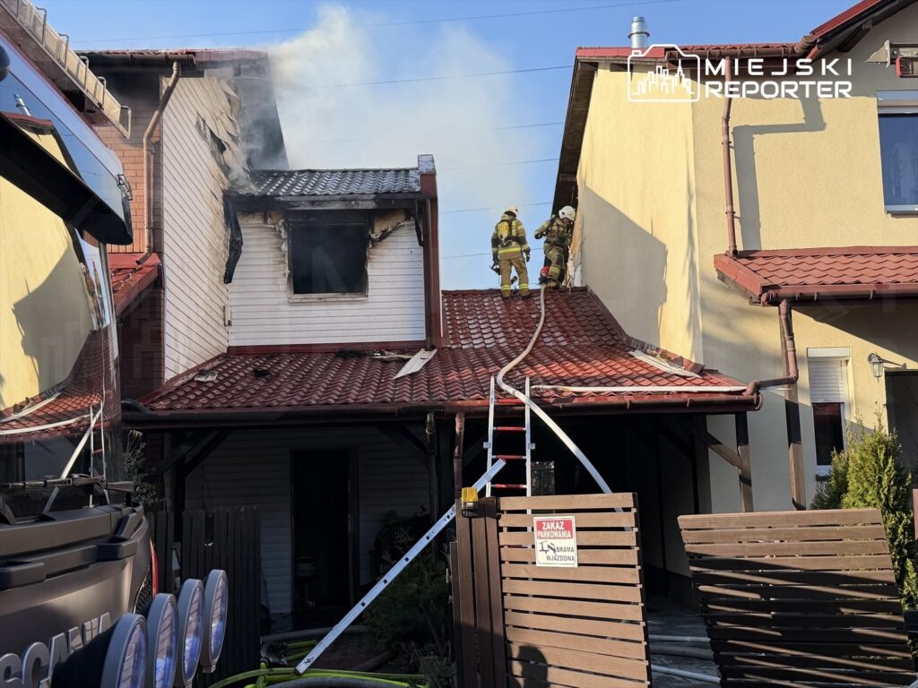 Dwóch strażaków na dachu domu, gaszących ogień wydobywający się z okna uszkodzonego pomieszczenia.
