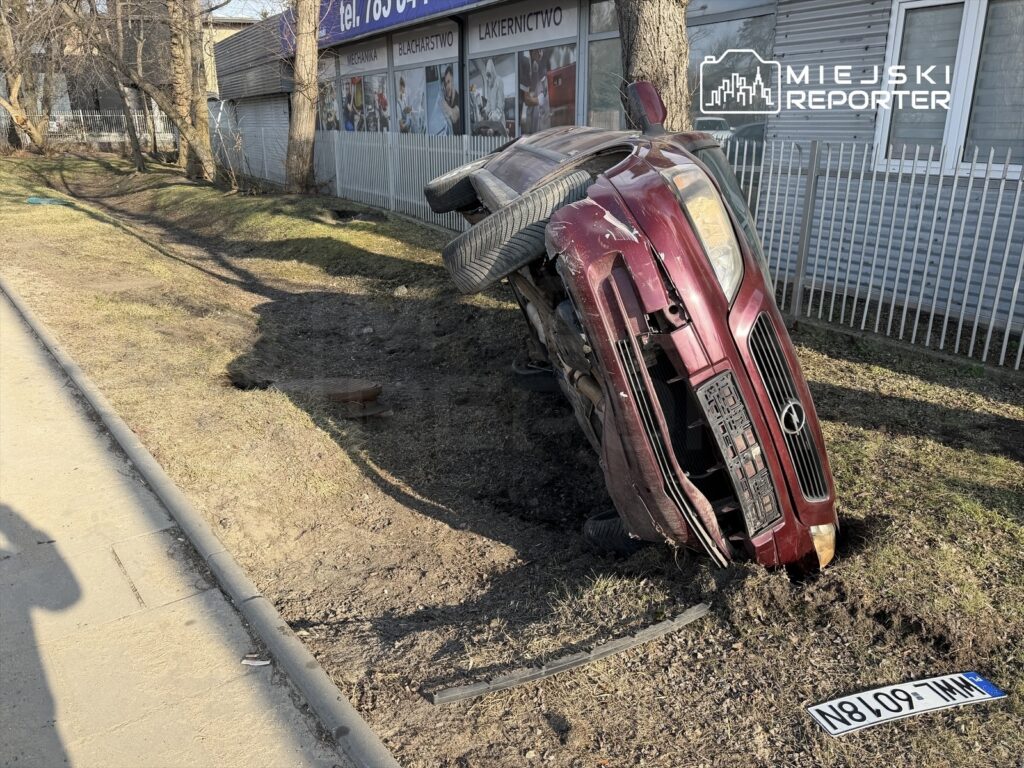 Czerwony samochód osobowy w pozycji na boku, utknął w rowie przy chodniku, obok ogrodzenia z białych paneli.
