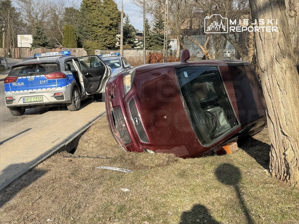 Czerwony samochód osobowy przewrócony na bok obok drogi, obok dwa radiowozy policyjne z włączonymi sygnałami świetlnymi.