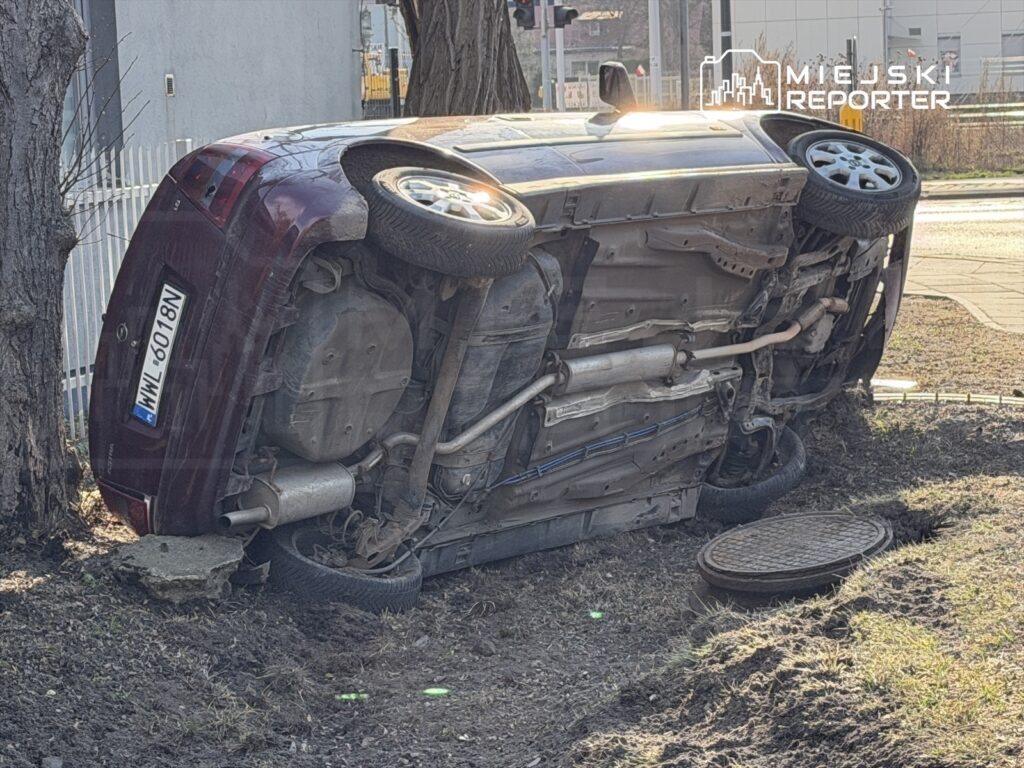 Czerwony samochód leży na dachu obok krawężnika, z widocznym uszkodzeniem nadwozia. Obok znajduje się studzienka kanalizacyjna.