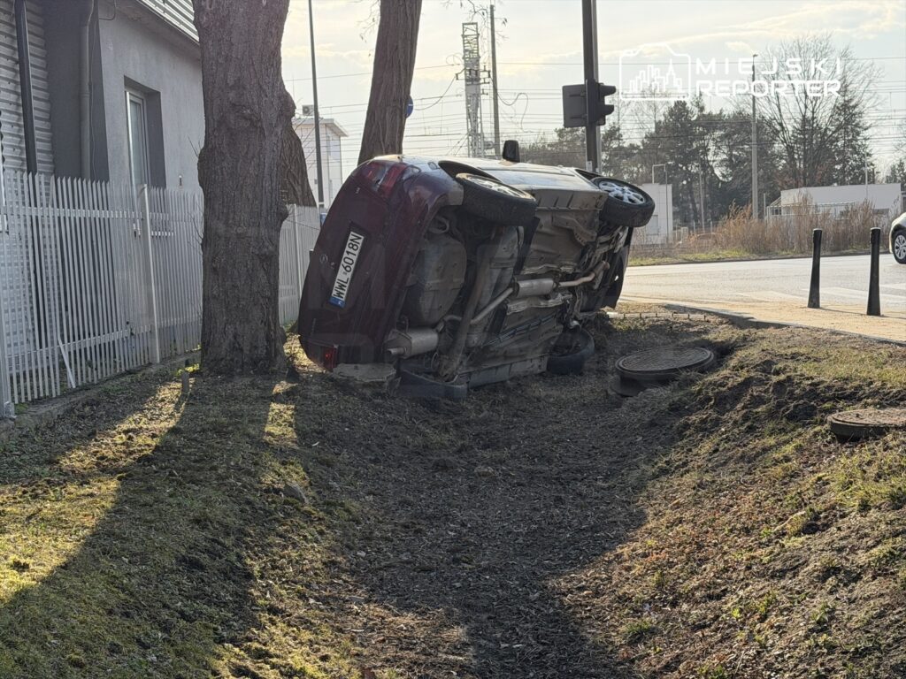 Czerwony samochód osobowy leży na boku w rowie przy drodze, obok drzewa i ogrodzenia.