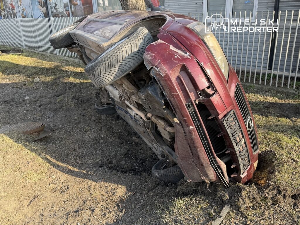 Czerwony samochód leży na boku w rowie, obok ogrodzenia, z widocznymi uszkodzeniami na karoserii.
