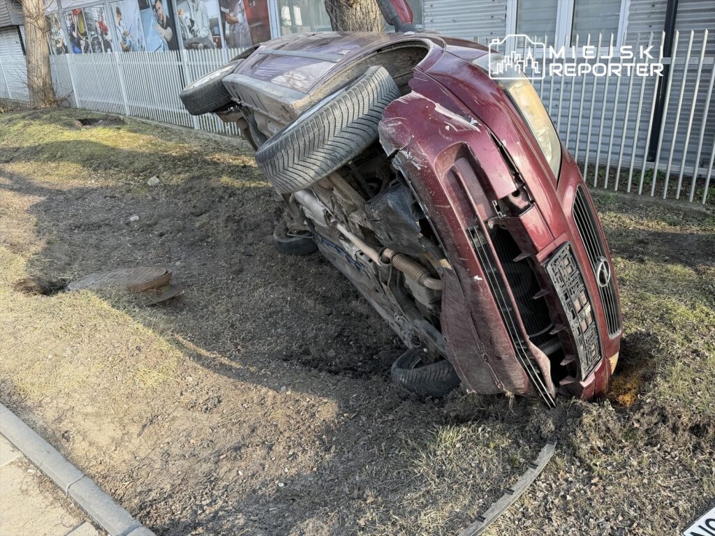 Czerwony samochód leży na boku w rowie obok chodnika, z jedną oponą uniesioną w górę.