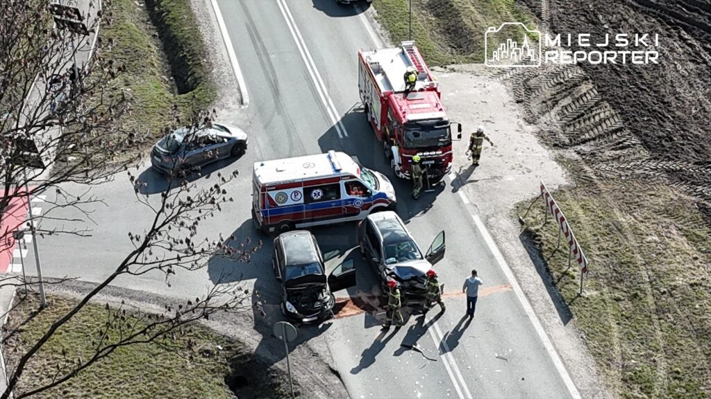 W miejscu wypadku drogowego stoją dwa rozbite samochody, obok nich zespół straży pożarnej i karetka.