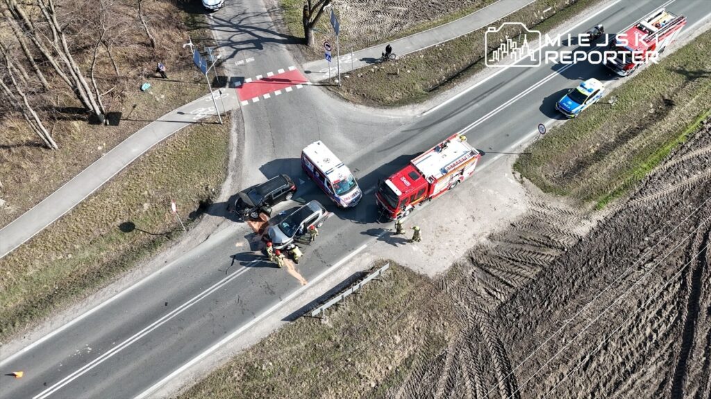 Na skrzyżowaniu dróg ratownicy medyczni udzielają pomocy poszkodowanym w wypadku drogowym, obok stoją dwa pojazdy służb ratunkowych.