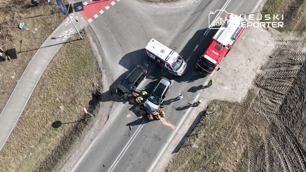 Na skrzyżowaniu dwóch dróg doszło do wypadku, w którym biorą udział dwa uszkodzone pojazdy. Ratownicy medyczni i strażacy udzielają pomocy poszkodowanym.