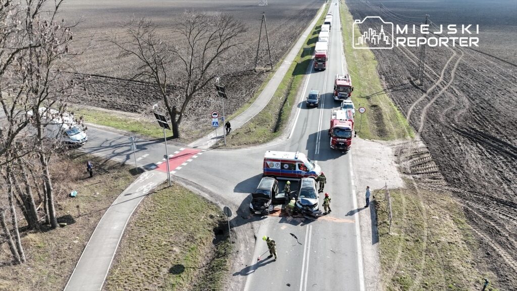 Dwa pojazdy osobowe zderzone na drodze, w tle strażacy i karetka, obok leży plama płynów eksploatacyjnych.