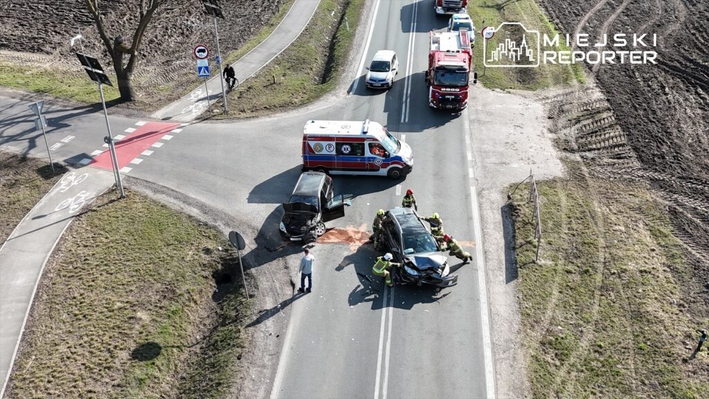 Na skrzyżowaniu dróg ratownicy medyczni oraz strażacy udzielają pomocy poszkodowanym w wypadku drogowym.