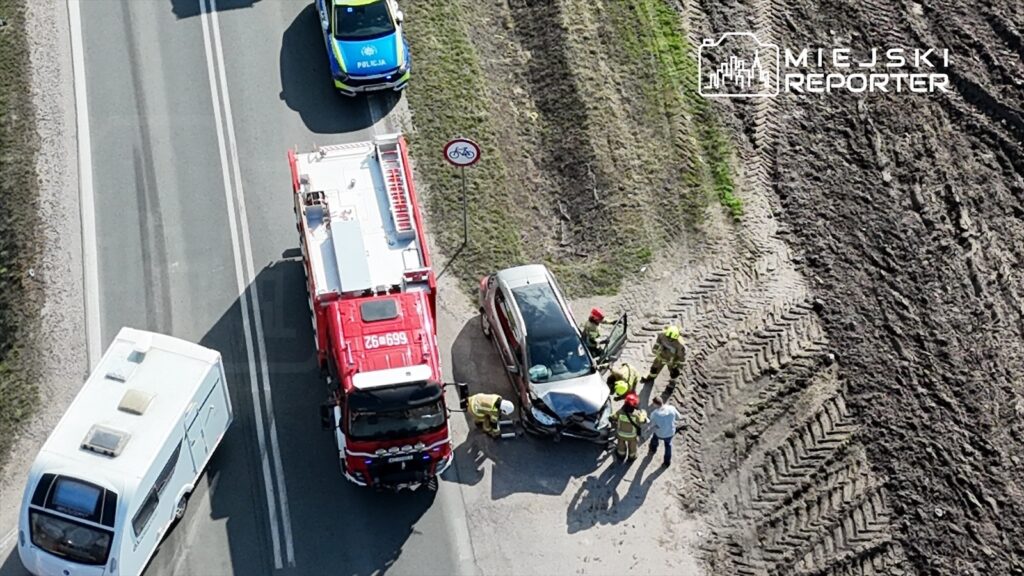 Wielu ratowników w mundurach pracuje nad rozbitym samochodem osobowym na drodze, obok stoi wóz strażacki.