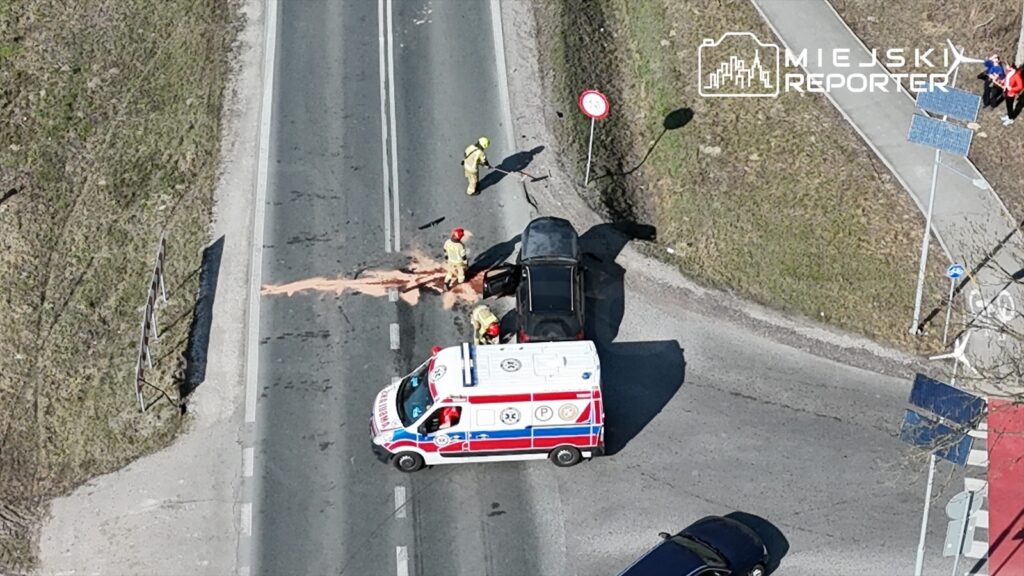Czterech strażaków w żółtych uniformach zabezpiecza miejsce wypadku na drodze, obok stoi ambulans.