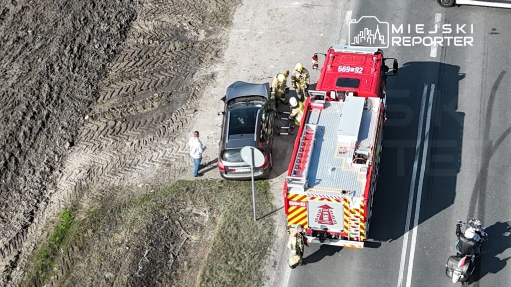 Strażacy w mundurach pracują przy uszkodzonym samochodzie osobowym na poboczu drogi, obok stoi mężczyzna.