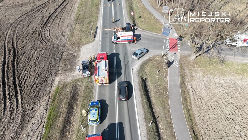 Na skrzyżowaniu drogi stoją pojazdy ratunkowe, w tym karetka i wóz strażacki, a obok pracują strażacy.
