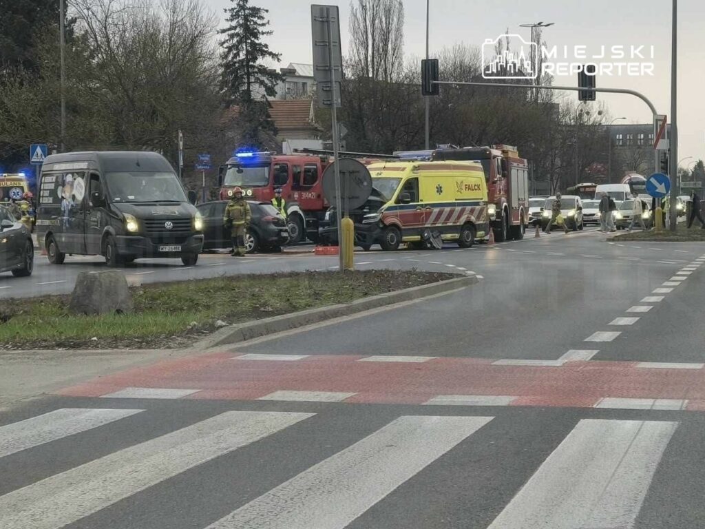 Na skrzyżowaniu widać pojazdy służb ratunkowych, w tym ambulans i wóz strażacki, oraz zablokowany ruch drogowy.