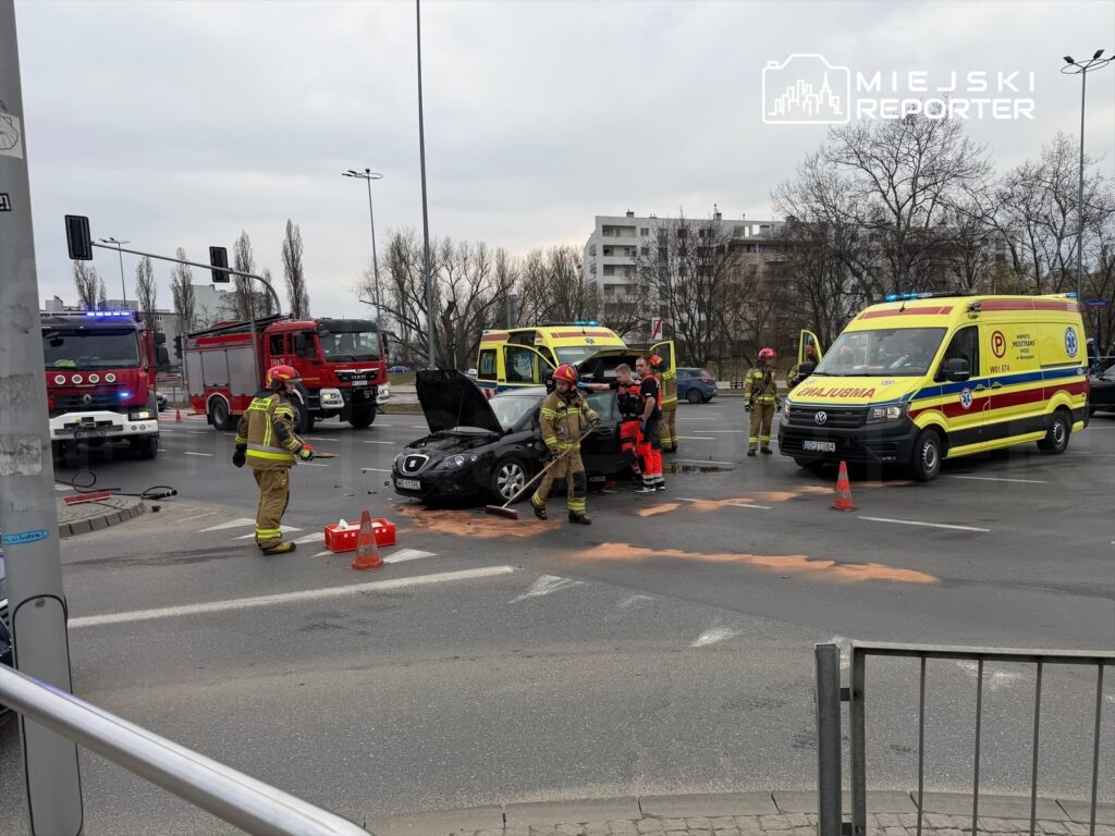 Strażacy i ratownicy medyczni pracują przy uszkodzonym samochodzie na skrzyżowaniu, obok pojazdów ratunkowych.