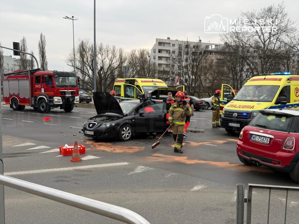 Strażacy sprzątają plamy oleju po wypadku drogowym, obok stoją dwa ambulanse i woz strażacki.