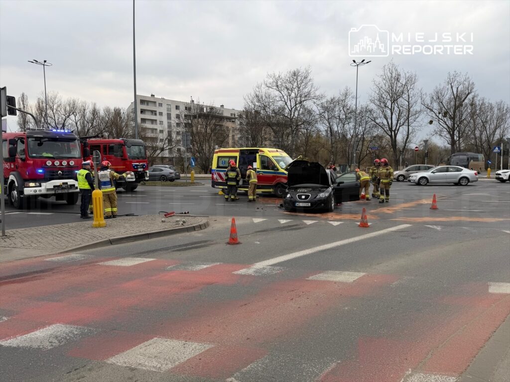 Na skrzyżowaniu pracują strażacy i policjanci przy uszkodzonym czarnym samochodzie, obok stoi karetka.